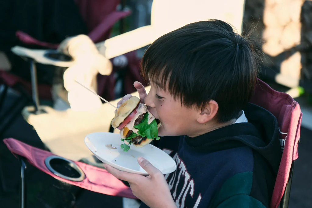 子どもが皿を手に持ち、ハンバーガーにかぶりついている。