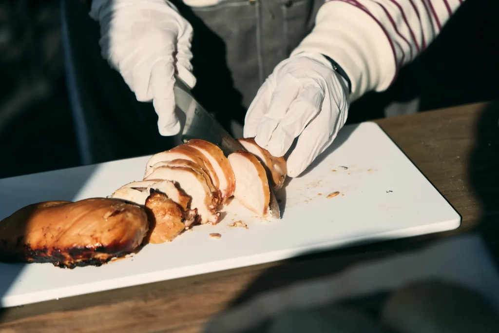 手袋をした人物が、白いまな板の上で焼いた鶏肉をナイフで切り分けている。