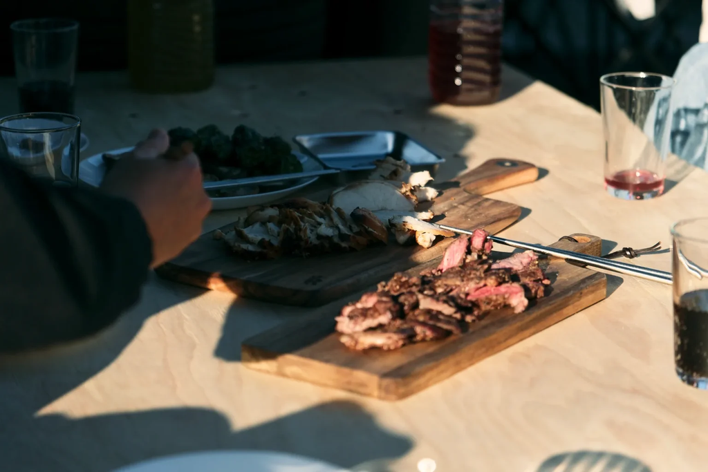 屋外のテーブルに、木製ボードに盛り付けた焼き肉や飲み物が並んでいる。